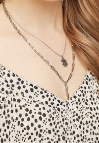 Stříbrný vrstvený náhrdelník s designem řetízku a kulatým přívěskem, nošený přes top s tečkovaným vzorem, který obsahuje černé a zlaté prvky.
