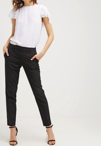 Bílá blůza s volánovými rukávy, kombinovaná s černými přiléhavými kalhotami a černými sandály na podpatku. Hladká tkanina, přiléhavý design, lehká.