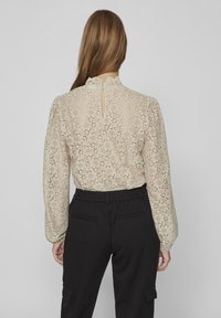 Donna con lunghi capelli castani che indossa una blusa in pizzo floreale beige con bottoni sulla schiena e pantaloni neri a vita alta, in piedi contro uno sfondo neutro.