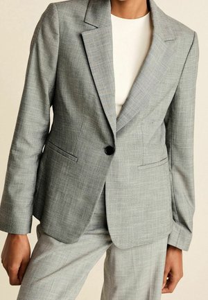 Personne portant un blazer à carreaux gris clair avec un seul bouton noir sur un haut blanc et un pantalon gris assorti.