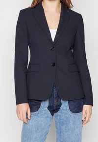 Granatowa marynarka o dopasowanym kroju, z klapą z wycięciem, jednorzędowym zapięciem, dwoma przednimi kieszeniami i czarnymi guzikami. W zestawieniu z niebieskimi dżinsami.