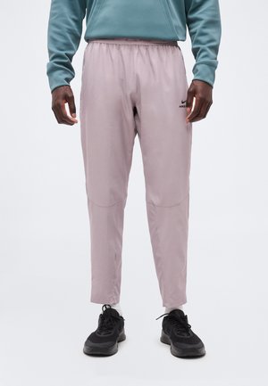 Muž oblečený v svetlo fialových Nike teplákoch, čiernych teniskách a oceánovej mikine, stojaci pred jednoduchým bielym pozadím.