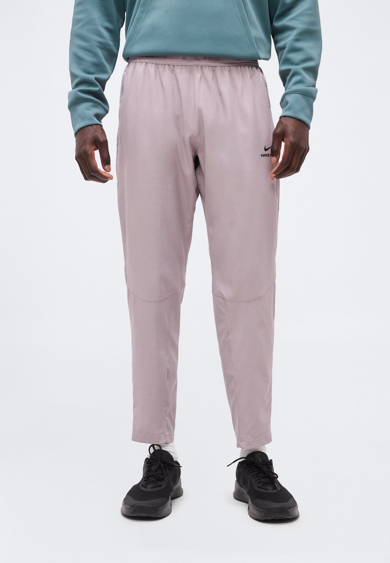 Άνδρας που φοράει ανοιχτό μωβ φόρμες Nike, μαύρα αθλητικά παπούτσια και τιρκουάζ φούτερ, στέκεται μπροστά σε απλό λευκό φόντο.