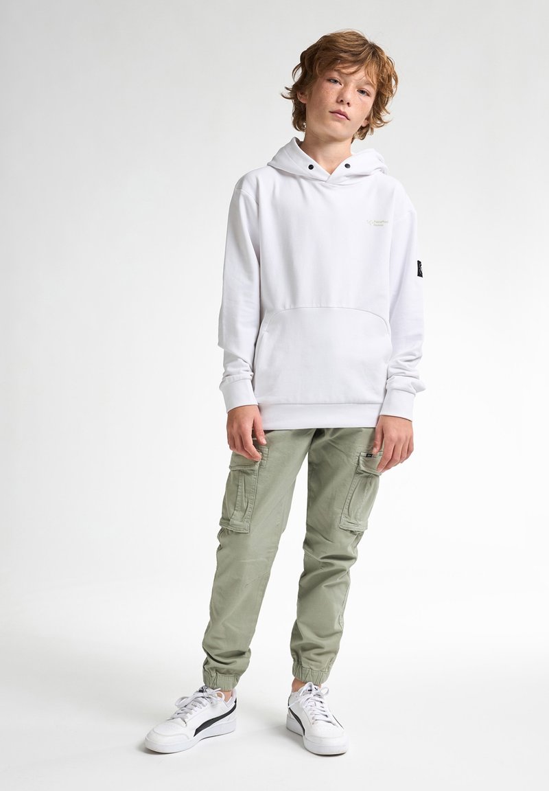 Felpa bianca con tasca frontale, abbinata a pantaloni cargo verde chiaro con tasche laterali e orli a gamba affusolata; sneakers bianche.