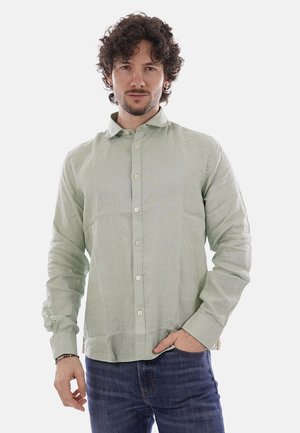 Uomo con capelli ricci indossa una camicia verde chiaro con bottoni e jeans blu, in piedi con una mano in tasca davanti a uno sfondo bianco.