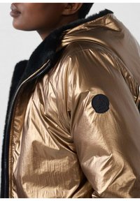Chaqueta acolchada en bronce metálico con capucha forrada de pelo negro, que presenta un parche de logo negro en la manga y una textura suave y brillante.