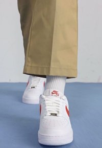 Fehér Nike Air Force 1 sneaker piros swoosh-szal, sima bőr anyagból, perforált orr résszel, bézs nadrággal és fehér zoknival párosítva.