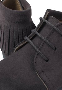 Zapatos de ante negro con puntera redondeada y detalle de flecos en el tobillo, con cordones planos negros y adornos de costura para un diseño adicional.