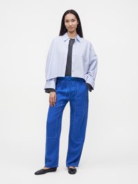 Jeune femme aux longs cheveux foncés portant une chemise courte bleu clair, un haut rayé, un pantalon bleu vif et des ballerines noires, debout devant un fond blanc.