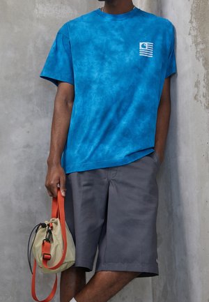 Personne portant un t-shirt tie-dye bleu et un short gris, tenant un petit sac beige avec une sangle orange, debout devant un mur texturé.