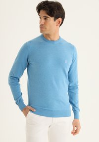 Pull bleu clair en tricot doux, avec un col rond, des manches longues, des poignets côtelés et un petit logo sur la poitrine.