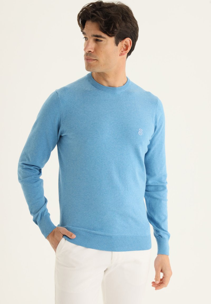 Pull bleu clair en tricot doux, avec un col rond, des manches longues, des poignets côtelés et un petit logo sur la poitrine.