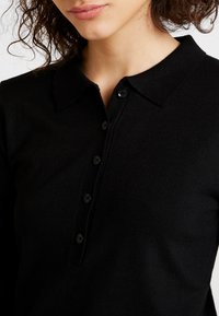 Zwart gebreid poloshirt met kraag, voorzien van een knoopsluiting en textuurstof. Close-up laat de stof en knoopdetails zien.