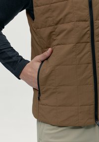 Hnědá prošívaná vesta se zipovou kapsou, vyrobená z lehkého materiálu. Má vysoký límec a je vrstvená přes tmavé tričko s dlouhým rukávem.