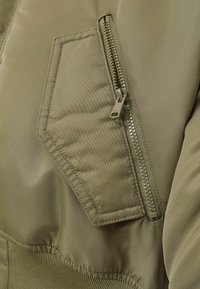 Nærbillede af en khaki stofjakke lomme med en skrå klap og en lodret lynlås ved siden af, detaljeret syning synlig.