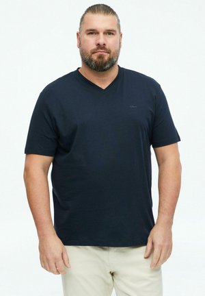 Man with short slicked-back hair and beard wearing a dark blue V-neck T-shirt and light beige pants, standing against a white background.