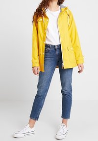 Gul vattentät regnjacka med huva, tryckknappar och framfickor, kombinerad med blå jeans och vita sneakers.