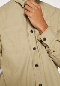 Camicia beige in tessuto testurizzato, con colletto, due tasche frontali e bottoni marroni lungo la zip e i polsini.