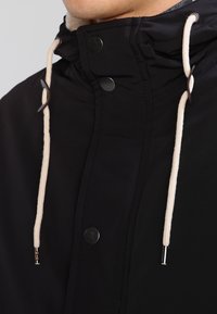 Svart jacka med hög krage, prydd med silverklikker och krämfärgade dragsnören. Slät textur med en elegant design och minimalistisk detaljering.