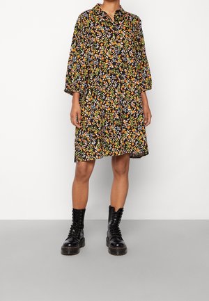 Frau trägt ein knielanges schwarzes Kleid mit orangefarbenen, weißen und grünen Blumenmustern und schwarze geschnürte Kampfstiefel.