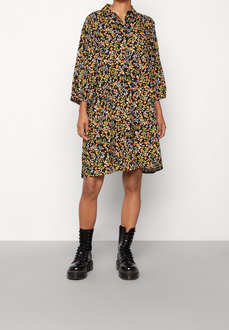 Femme portant une robe noire fleurie longueur genou avec des motifs orange, blanc et vert et des bottes de combat noires à lacets.