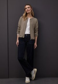 Beige gesteppte Bomberjacke, darunter ein weißes Hemd, kombiniert mit dunkelblauen Jeans und hellen Sneakers; stehend vor einem dunklen Hintergrund.