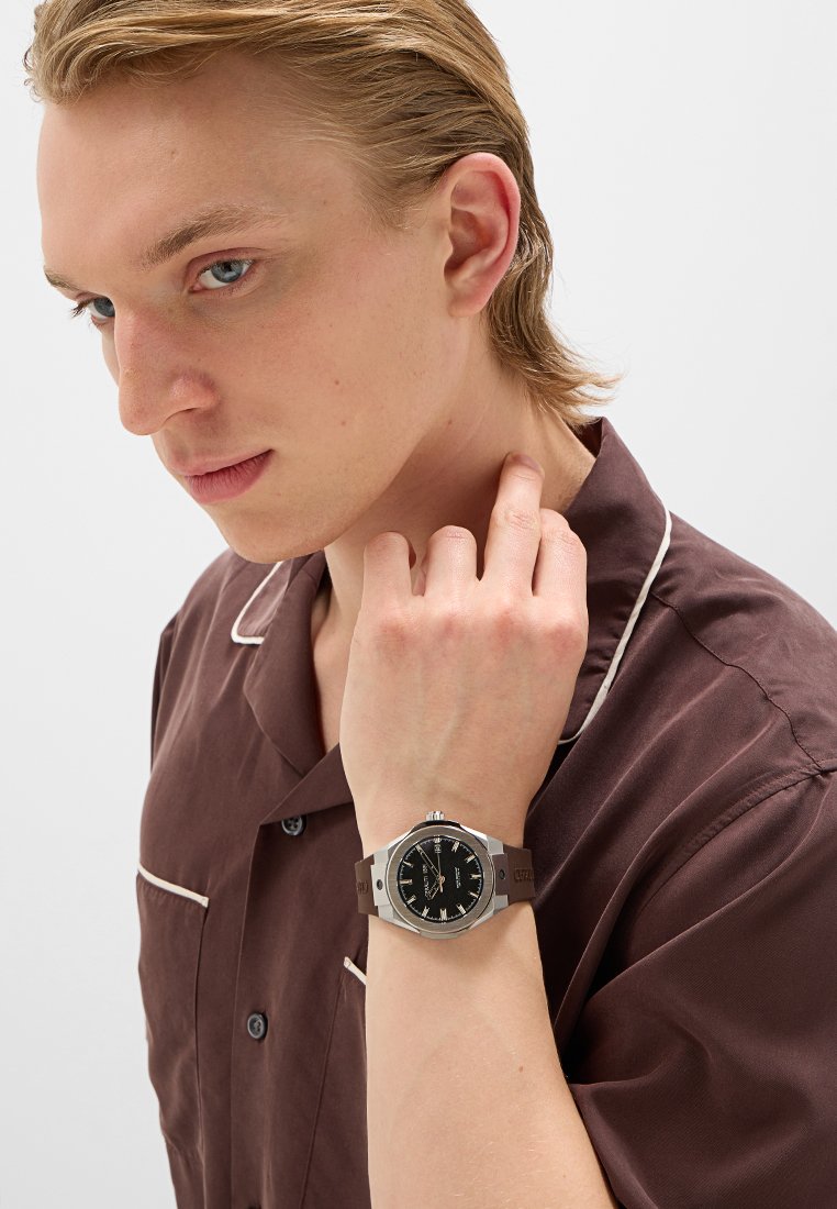 Montre pour homme avec un boîtier en métal argenté, un cadran noir et un bracelet marron. La montre présente une forme ronde et des index horaires clairs.