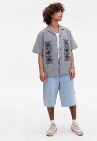 Camisa de manga curta em padrão gingham preto e branco com padrões bordados a preto, combinada com calções de ganga azul claro e ténis bege.