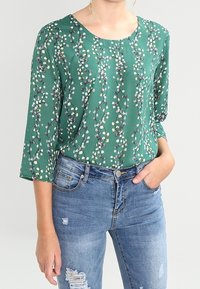 Grüne Blumenbluse mit rundem Halsausschnitt und drei Viertelärmeln, versehen mit einem Muster aus kleinen Blumen auf einem glatten Stoff. Kombiniert mit abgetragenem Denim.