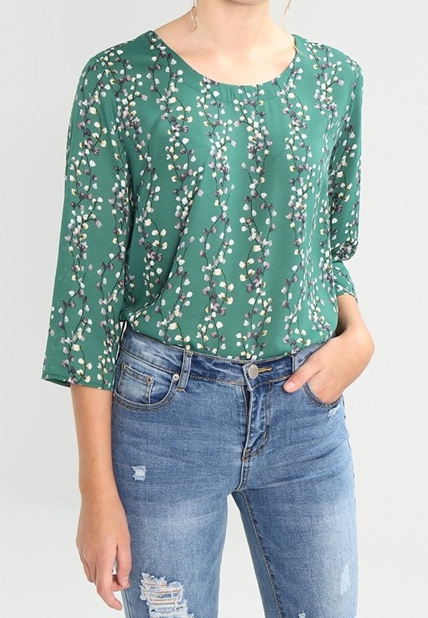 Grüne Blumenbluse mit rundem Halsausschnitt und drei Viertelärmeln, versehen mit einem Muster aus kleinen Blumen auf einem glatten Stoff. Kombiniert mit abgetragenem Denim.
