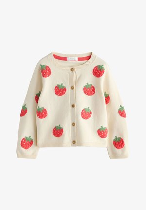 Jeune enfant aux cheveux bouclés portant un cardigan blanc avec des fraises et un jean bleu, tenant des fleurs sauvages dans les poches à l'extérieur par une journée ensoleillée.