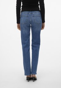 Jeans denim gelap dengan potongan kaki lurus, dilengkapi dengan saku belakang dan sabuk yang terlihat. Dipadukan dengan sepatu hak tinggi hitam.