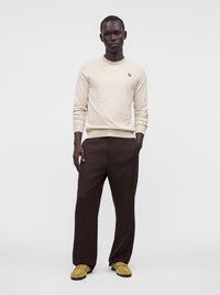 Homme portant une chemise beige à manches longues, un pantalon marron foncé et des chaussures jaune moutarde, debout devant un fond blanc uni.
