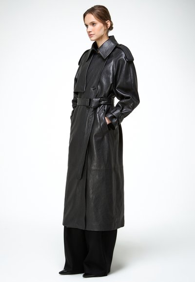 Vrouw draagt een lange zwarte leren trenchcoat met een riem, brede revers en zijzakken, gecombineerd met brede zwarte broek en zwarte schoenen.