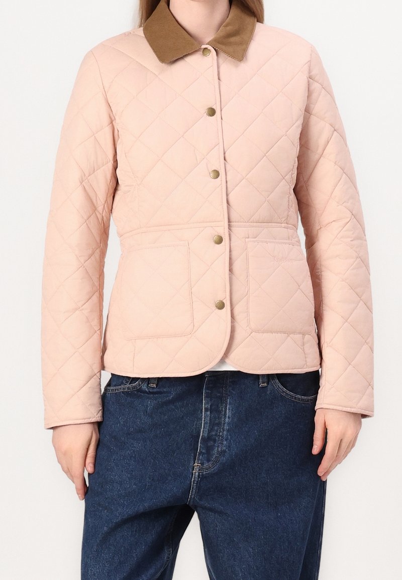 Veste matelassée rose clair avec col marron, fermeture par boutons et poches avant, portée avec un jean bleu foncé.