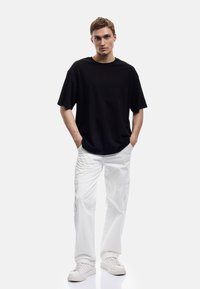 Sort oversized t-shirt i bomuld, parret med hvide cargobukser med lommer. Modellen bærer hvide sneakers og står oprejst foran en ensfarvet baggrund.