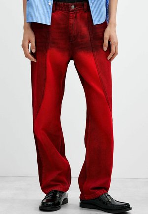 Person iført røde vidde-jeans med sorte loafers og en blåstribet skjorte, stående mod en ensfarvet baggrund.
