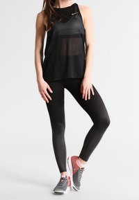 Mujer vistiendo una blusa sin mangas de malla negra, leggings texturizados negros y zapatillas deportivas grises y coral, de pie con una pierna cruzada.