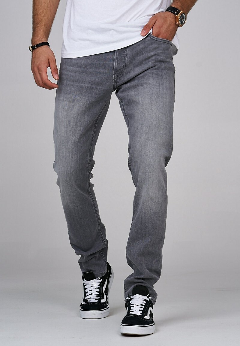 Jeans in denim grigio con una vestibilità slim, caratterizzati da un classico design a cinque tasche. Indossati con sneakers in bianco e nero, evidenziando uno stile casual.