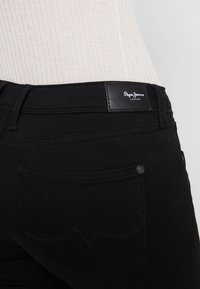 Vaqueros de denim negro con una textura suave, que cuentan con un parche de logo de cuero en la cintura y costuras decorativas en los bolsillos traseros.