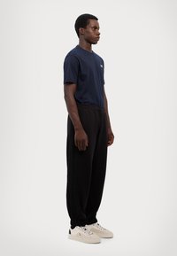 Navy blue cotton t-shirt, black track pants with side stripes, and light sneakers with black accents. Model stands in profile.
