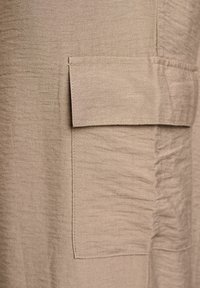 Beige látka s texturovaným povrchem, se širokou náprsní kapsou a jemným zvrásněním, což zdůrazňuje uvolněný a neformální design.