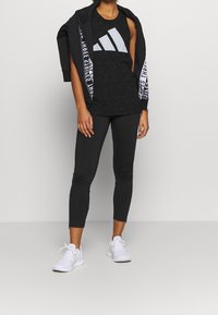 Canotta nera con grande logo grigio adidas, abbinata a leggings neri con dettagli zip alle caviglie. Modella in sneakers bianche.