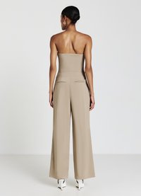 Combinaison sans manches beige avec jambes larges, design ajusté et poches arrière cousues. Portée avec des chaussures à talons blancs.