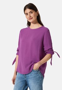 Lila texturerad långärmad skjorta med knutna manchetter, rund halsringning och en lös passform, kombinerad med ljusblå jeans.