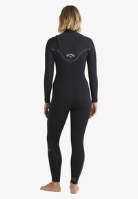 Person, der barfuß einen schwarzen Neoprenanzug mit langen Ärmeln trägt, sichtbares Billabong-Logo auf dem oberen Rücken und den unteren Beinen.
