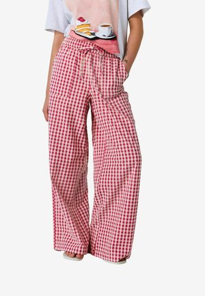 Personne portant un pantalon large à carreaux rouges et blancs avec une taille à cordon et un t-shirt gris avec un imprimé gâteau et café.