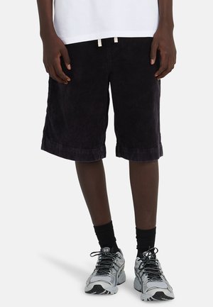 Person trägt schwarze kniehohe Shorts, weißes Oberteil, schwarze Socken und graue Sportschuhe und steht vor weißem Hintergrund.