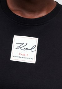 Černé bavlněné tričko s kulatým výstřihem, se čtvercovou bílou etiketou s černým a červeným textem: "Karl," "PARIS," a "21 RUE SAINT-GUILLAUME."