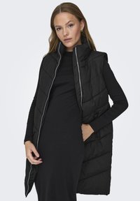Veste rembourrée noire avec un col haut, dotée d'une fermeture éclair et d'un motif matelassé, portée sur une robe noire à manches longues.